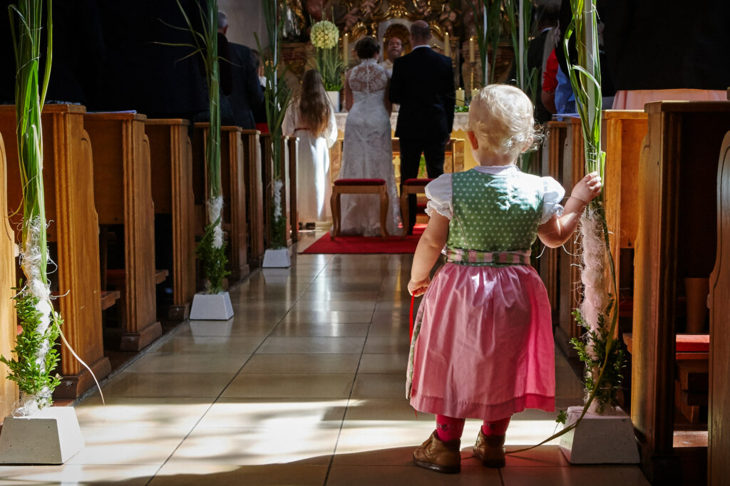 Hochzeitsbilder Ideen: Ein kleines Mädchen im Dirndl steht während der kirchlichen Trauung im Gang