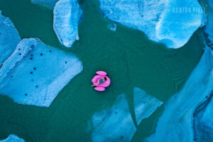Drohnenaufnahme in Island: Eine Frau liegt auf einem pinkfarbenen Schwimmtier-Pelikan im Badeanzug zwischen Eisschollen im türkisfarbenen Wasser